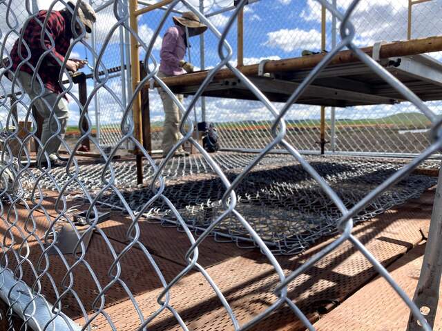 Joe and Daphne installing chain link fencing to cover holes in platform. The ends of the fencing are fixed with tension bars that are either drilled into the platform or secured with u-nails.