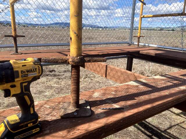 We moved a 2x12 plank from the edge of the platform to the middle so that we had more area to secure the scaffolding foot. The feet were drilled into the platforms and the scaffolding was tied down with a chain and turnbuckle attached to an eyebolt screwed into the platform (not pictured). The holes in the platform were later covered by chain link fencing.